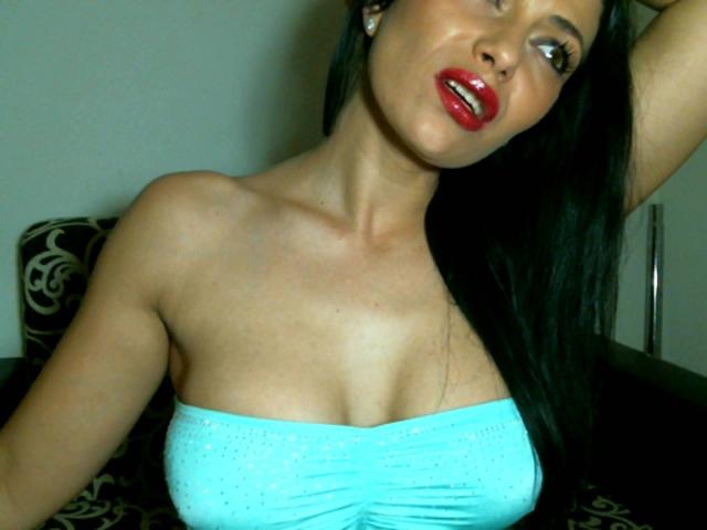 DeeLicious with red lipstick, black hair, and a blue strapless top showing her tits, raising an arm, and looking to the side. Her skin is tan and she's sexy.
