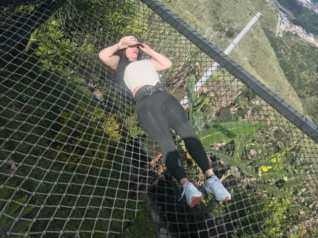 KinkyVane hanging from a black chain-link fence, wearing a white top, black leggings, belt, and white sneakers, with her hand shielding her eyes from sunlight.