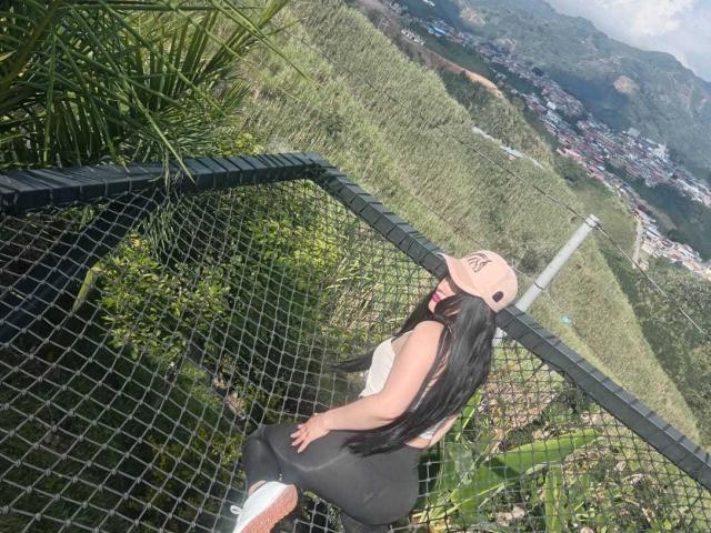 KinkyVane with long black hair, wearing a pink cap, white tank top, and black leggings, standing on a green, grassy hill, looking over a black chain-link fence.