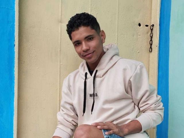 BrawnLove, short curly black hair, light brown skin, white hoodie, blue jeans, slight smile, hand on knee, ear piercing, blue and yellow wall, chain on wall, relaxed.