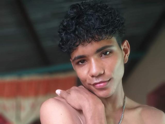 BrawnLove has curly black hair, medium brown skin, and a slight smile, with his hand on his shoulder, wearing a chain necklace, looking at the camera.