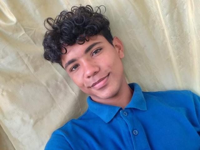 BrawnLove with curly black hair, brown eyes, and a slight smile, wearing a blue polo shirt, standing against a beige curtain. His ass is not visible.