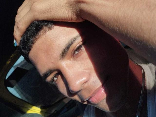 BrawnLove's face, shadowed on the right, with short black hair, tan skin, and a hand gently touching his head, showing a slight, relaxed smile. Sunlight highlights his forehead.