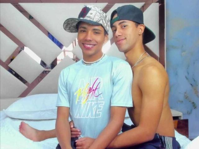 Two young guys sitting on a bed, BrawnLove on the left wearing a "Yellow Like" shirt and cap, BrawnLove on the right shirtless with a cap, both smiling.