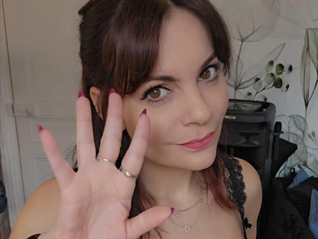 Close-up photo of JuneLaFrenchie, brown-haired woman with pink lipstick, black lace top, raised hand showing red-painted nails, rings, and necklace. Seductive smile, sexual tension, soft lighting.