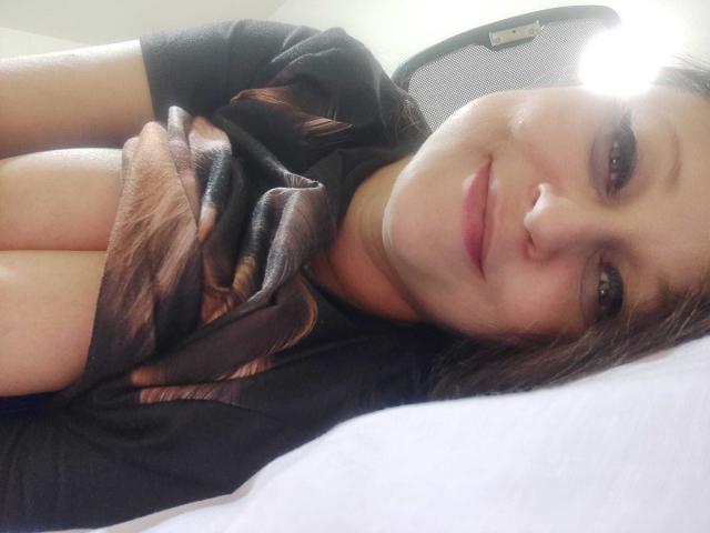 KissAndTits with brown hair, smiling, lying down, black shirt, exposing her large, soft, bare tits, slightly covered by a brown patterned scarf, on a white pillow.