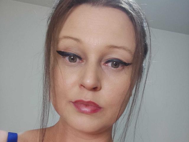 KissAndTits tiene ojos marrones con delineador negro grueso, lápiz labial rosado-rojizo y cabello castaño recogido en un moño suelto, y lleva una correa azul. Mira directamente a la cámara..