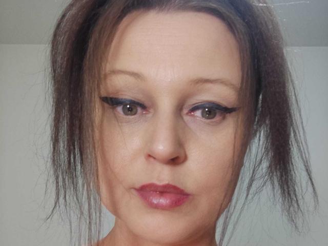 KissAndTits has brown eyes, black eyeliner, and pink lipstick. She has messy brown hair in a ponytail, a fair skin tone, and a serious expression.