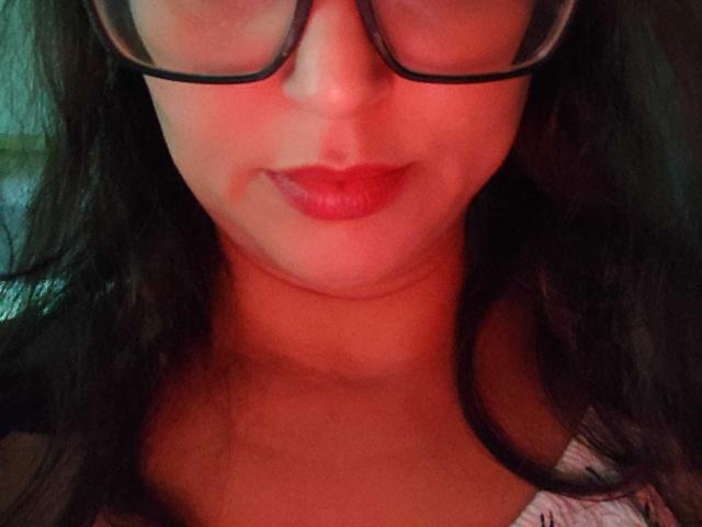 KissAndTits mit roten Lippen, dunklen, welligen Haaren und einer schwarzen Brille, die ein leichtes Lächeln zeigt. Ihre Brüste sind nicht sichtbar, aber ihr Kinn und der obere Brustbereich sind zu sehen..