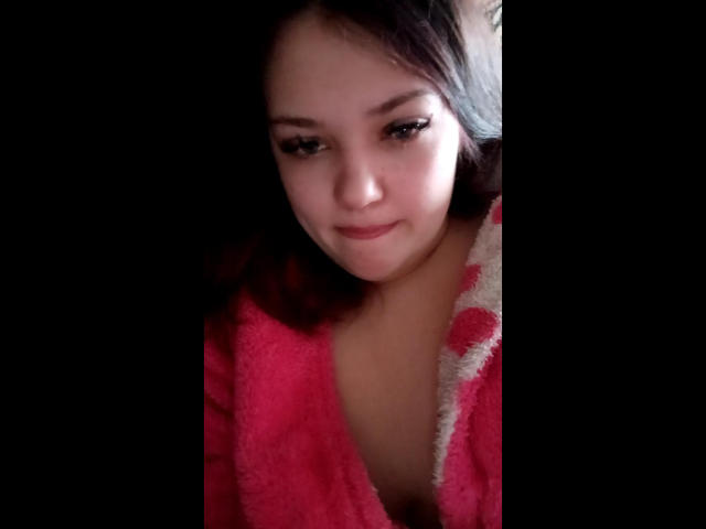 MalyaS with fair skin and dark hair, wearing a red, fluffy robe, looking down with a subtle smile, showing her small, perky tits. Dark background.