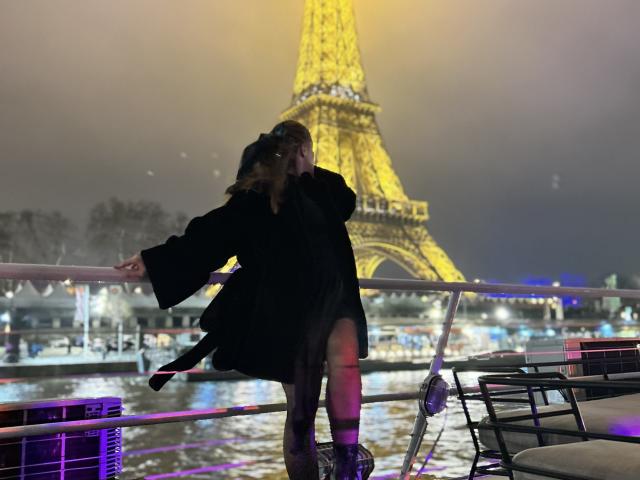 CamillaSweet, in a black coat, leans against a railing, kissing someone, with the Eiffel Tower glowing yellow in the night. Water reflections, chairs, and purple lights in foreground.