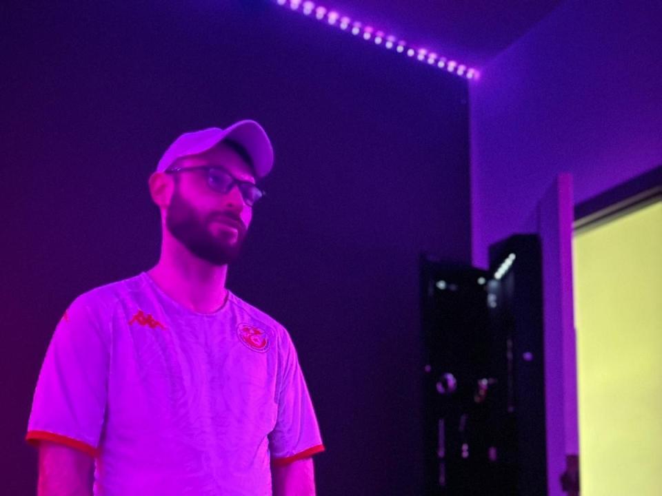 Un uomo di nome BearSoumis, con occhiali e barba, che indossa un berretto bianco e una camicia illuminata di viola, in piedi in una stanza con strisce LED viola. La camicia ha bordi arancioni..