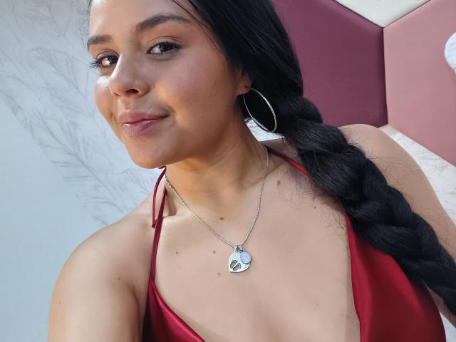 ArielleWilde with long black braided hair, red halter top, small silver necklace with heart, and hoop earrings, smiling slightly, showing her cute tits and cute ass.