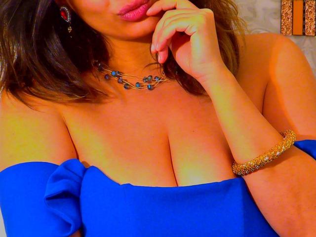 ScarletOra, avec une poitrine de taille moyenne, porte une robe bleue à une épaule, un collier bleu, un bracelet doré et des boucles d'oreilles rouges, et elle touche ses lèvres avec son doigt..