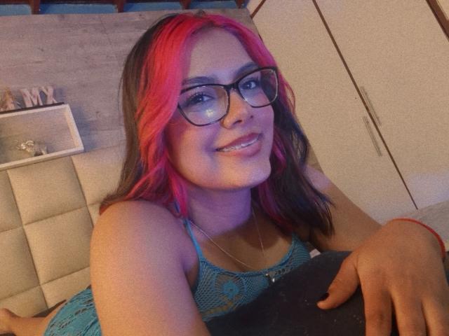EmilyRoberts with pink and black hair, glasses, smiling, wearing a blue lace top, lying on a bed, black thigh-highs, red bracelet, cute, attractive, and sexy.