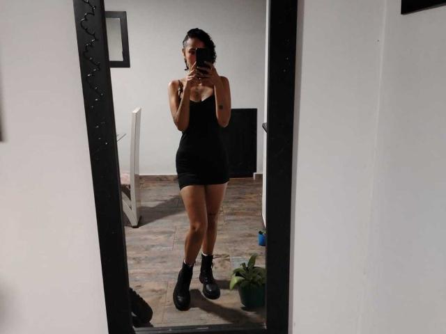 SilvanaXOwen in a black, tight, strapless dress with a short skirt, holding a phone, standing in front of a mirror, wearing black, chunky, ankle boots, and white socks.