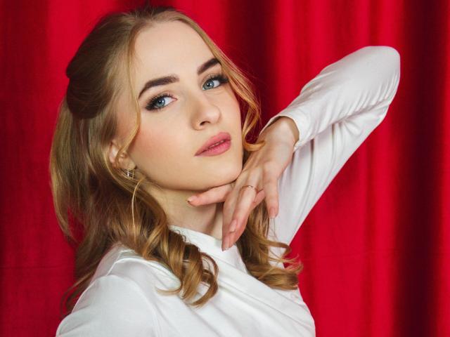 EvaDaly with blonde hair in pigtails, blue eyes, and white shirt, hand on neck, red background, wearing gold earrings, looking up with slightly parted pink lips.