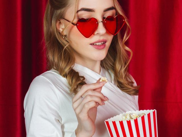 EvaDaly with heart-shaped red sunglasses, blonde curls, white shirt, holding popcorn in red and white striped container, eating, smiling, pink lipstick, red curtain.