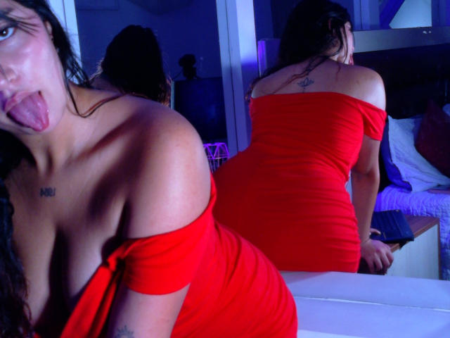 MelissaSmithers in a red off-shoulder dress, showing her tits and ass, with tongue out, mirror reflection, tattoo on back, and blue lighting, on a bed.