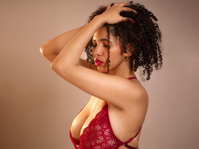 ZoeJonees with curly black hair, red lace bra, and red lipstick, arms raised, hands in hair, showing her big tits, light brown skin, and a sexy, confident expression.