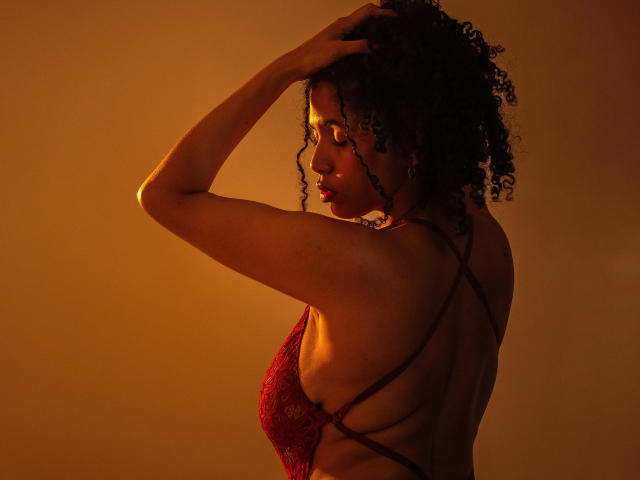 ZoeJonees with curly hair, wearing a red lace bra, has her arms up, back to camera, showing her ass and sideboob, in warm orange lighting.