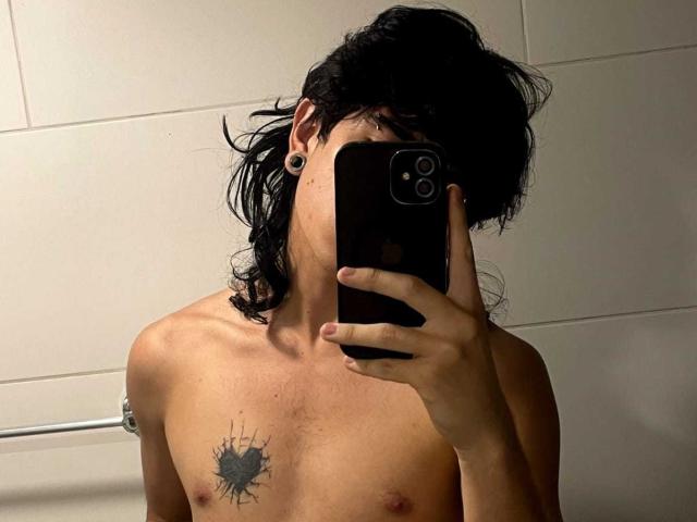 AxellKush with messy black hair, holding a phone covering their face, shirtless with small tits, a black splatter tattoo on the chest, and black ear gauges, in a bathroom.