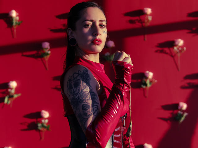 LusstCruella with tattoos on her arm, wearing red latex gloves and dress, dark hair, black ear plugs, red lipstick, standing against red wall with roses, sunlight casting shadows.