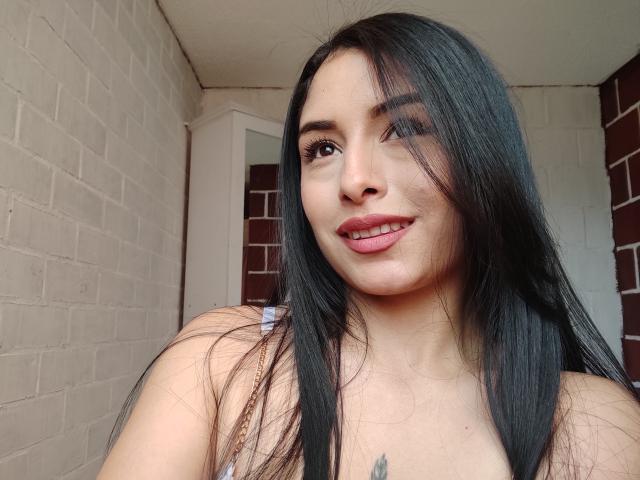 EmilyVv with long black hair, brown eyes, pink lipstick, smiling, showing her tits, has a small tattoo on her chest, standing against a white brick wall.