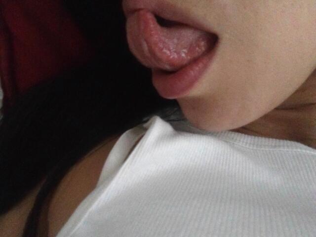 SeleneBrigth's tongue is out, licking her upper lip with glossy pink lips and a slight wet sheen. She's wearing a white ribbed tank top, showing her bare shoulders.