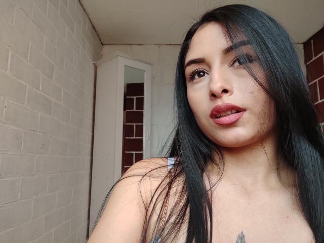 EmilyVv, con cabello negro largo, labios rosados y un tatuaje cerca del pecho, mirando a la cámara con la boca ligeramente abierta. Sus senos son parcialmente visibles..