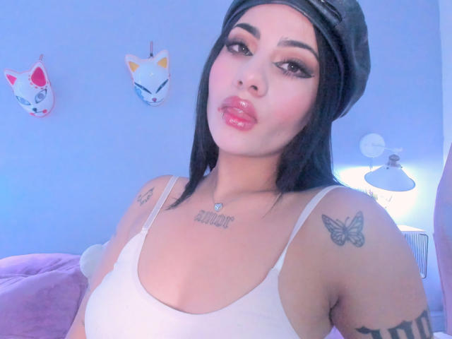 NatallieBlue with glossy pink lips, black beanie, white tank top showing her boobs, tattoos on shoulders and chest, blue-tinted bedroom, dark hair, makeup, and butterfly tattoos.