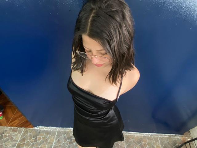 ShyVibes, avec ses cheveux noirs mi-longs, portant des lunettes et une robe en satin noir, debout devant un mur bleu, regardant vers le bas avec une expression timide, montrant ses petites seins..