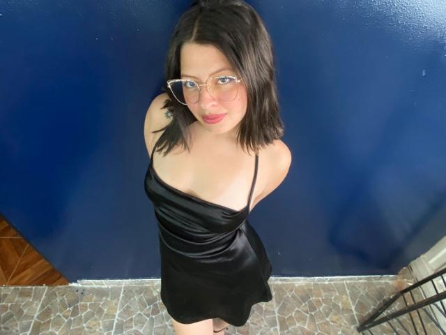 ShyVibes with black dress, glasses, and red lipstick, looking up, has small tits, black hair, and a cute face, standing against a blue wall, on a tiled floor.