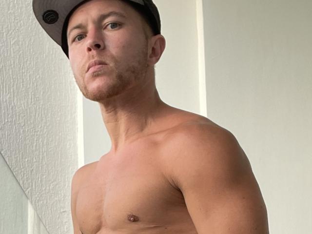 AaronSky, shirtless, wearing a dark cap, with a serious expression, light skin, toned muscles, brown eyes, and a slight beard, standing in front of a white wall.