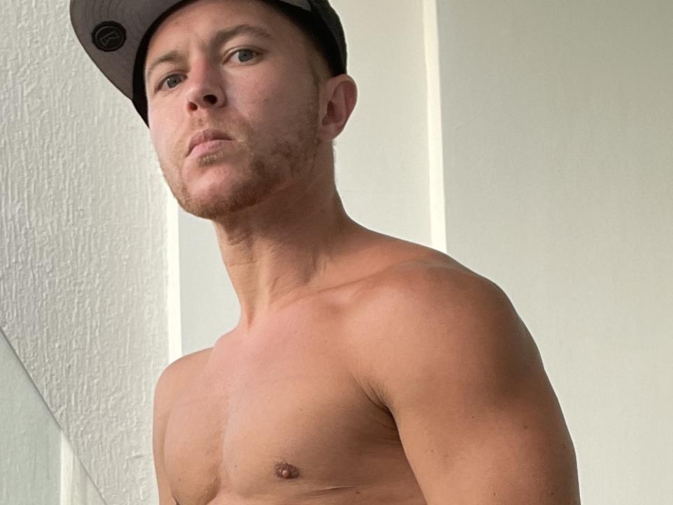 AaronSky, torse nu, portant une casquette sombre, avec une expression sérieuse, peau claire, muscles toniques, yeux marrons et une légère barbe, debout devant un mur blanc..