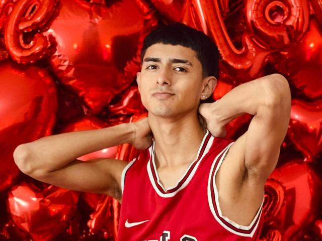 NathanMaxime, met zijn armen achter zijn hoofd, draagt een rood basketbalshirt, waardoor zijn gespierde armen en korte zwarte haar te zien zijn. Hij kijkt zelfverzekerd met een lichte grijns. Rode ballonnen op de achtergrond..