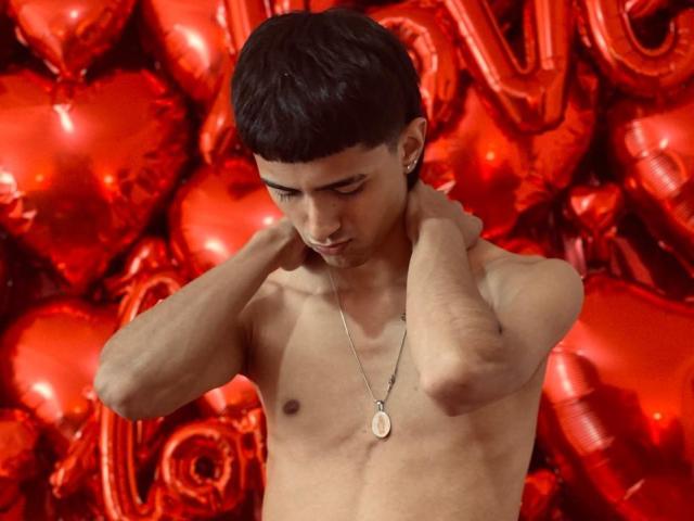 NathanMaxime, shirtless with short black hair, small penis, tan skin, necklace, looking down, hands on neck, surrounded by red heart-shaped balloons.