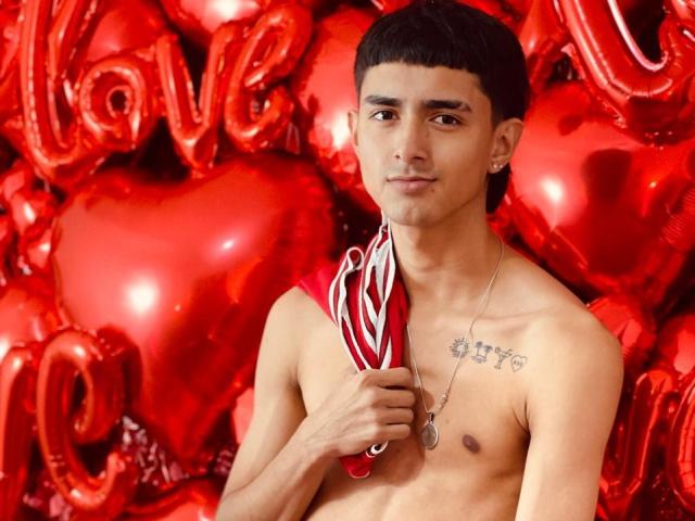 NathanMaxime, shirtless with a tattoo on his chest, holding a red and white striped cloth, surrounded by shiny red hearts and love balloons. He's got short black hair.