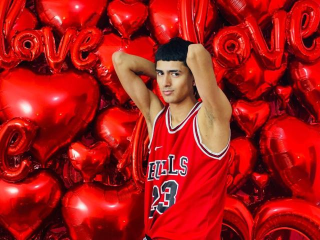 NathanMaxime staat met zijn handen in zijn nek, gekleed in een rood basketbalshirt, met rode hartjes en ballonnen met de tekst "LOVE" op de achtergrond. Hij heeft een gemiddeld postuur, donker haar en een serieuze blik..