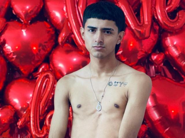 NathanMaxime, zonder shirt, met een slank postuur, een klein borstgedeelte, kort donker haar, tatoeages op de linkerborst, draagt een ketting, staat voor hartvormige rode ballonnen en kijkt naar de camera..
