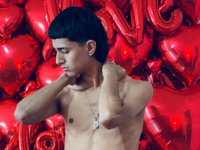 NathanMaxime, ohne Hemd, mit kurzen schwarzen Haaren, Ohrringe, Halskette, die sich am Hals festhaltend, nach unten schauend, im Hintergrund rote, herzförmige Ballons..