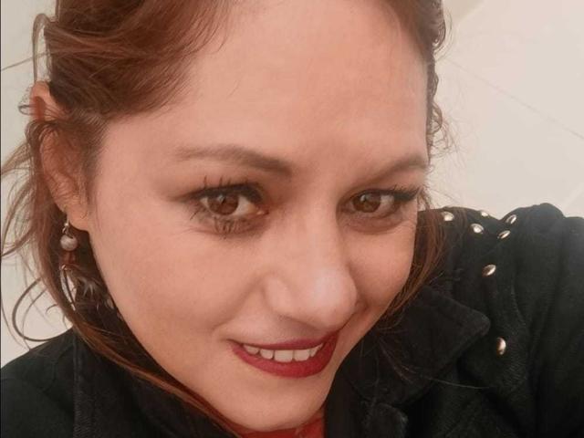 NatashaChaus has brown eyes, red lipstick, and a slight smile. She's wearing a black jacket with gold buttons and pearl earrings. Her brown hair is partially tied back.