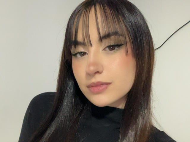 MiaHarperr tem cabelo castanho escuro liso com franja, delineador preto e lábios rosa. Ela está vestindo uma blusa preta, olhando ligeiramente para baixo com um sorriso discreto..