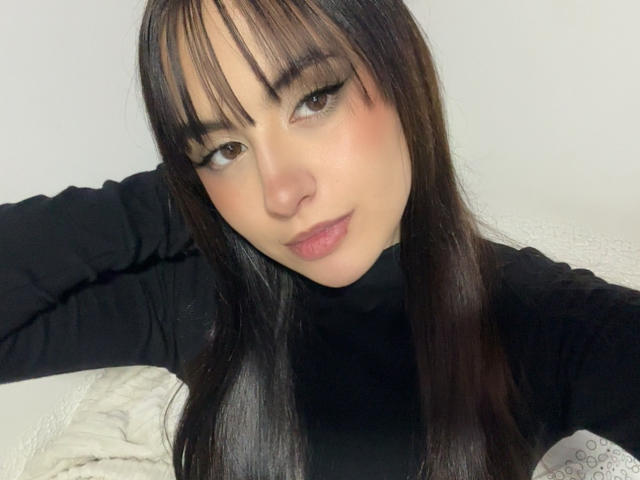 MiaHarperr has dark brown hair with bangs, black eyeliner, and is wearing a black turtleneck. She's smiling slightly, looking at the camera with brown eyes.