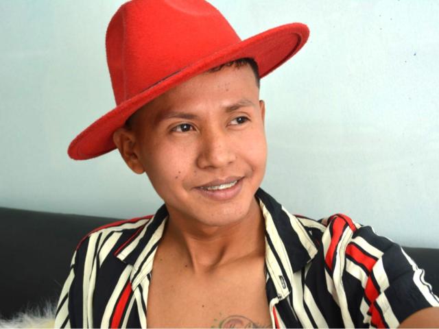 NathanHoper, wearing a red hat, black and white striped shirt, smiling, has a small tattoo on his chest, short brown hair, and light brown skin.