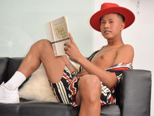 NathanHoper, shirtless, wearing a red hat, striped shorts, white socks, and white sneakers, sits on a black couch, reading a book, with a fluffy white pillow.