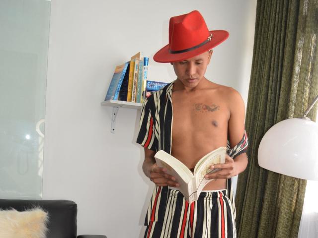 NathanHoper, shirtless with striped pants, red hat, and tattoos, reads a book. His brown skin, small chest, and flaccid ass are visible. Green curtains and white wall in background.