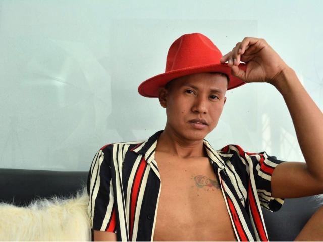 NathanHoper wearing a red hat, black and white striped shirt, and no shirt, showing a small tattoo on his chest, sitting on a black sofa with a white pillow.