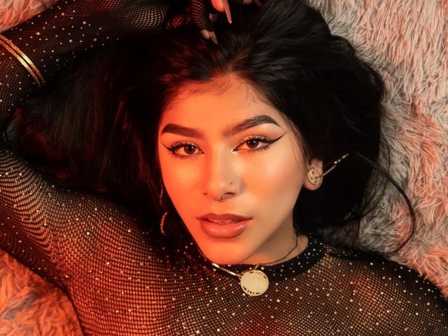 ZoeCarlter with dark hair, wearing a sparkly black mesh top, has a nose ring, winged eyeliner, and a gold necklace with a circular pendant, lying on a furry surface.