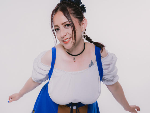CatalinaMonyque with huge tits in a blue dress, white puffy sleeves, black choker, and braid, leaning forward smiling, showing a small tattoo on her chest.